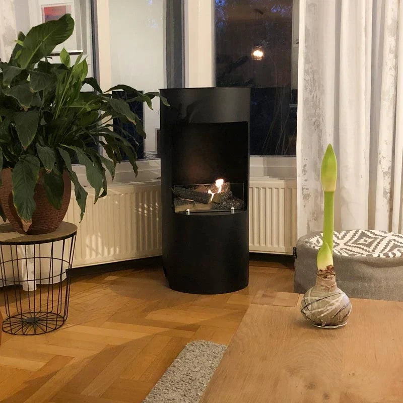 Montgomery black bioethanol fireplace lit in a corner nook with a large plant and evening lighting